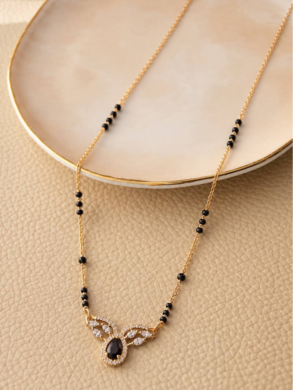 Mangalsutra with black beads and CZ pendant laid flat on a ceramic plate against a beige surface