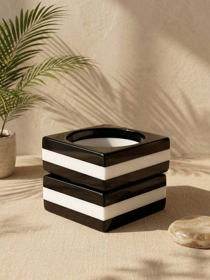 Single black and white striped resin square bangle resting on a linen surface beside a palm leaf