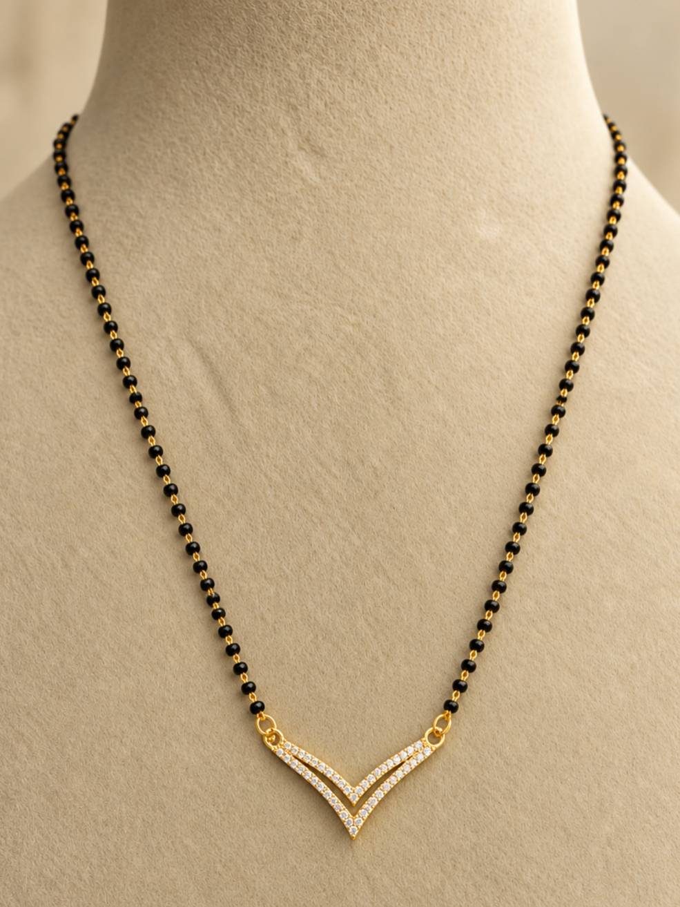 Gold-plated double V CZ mangalsutra displayed on a cream jewellery bust, showing full black bead chain