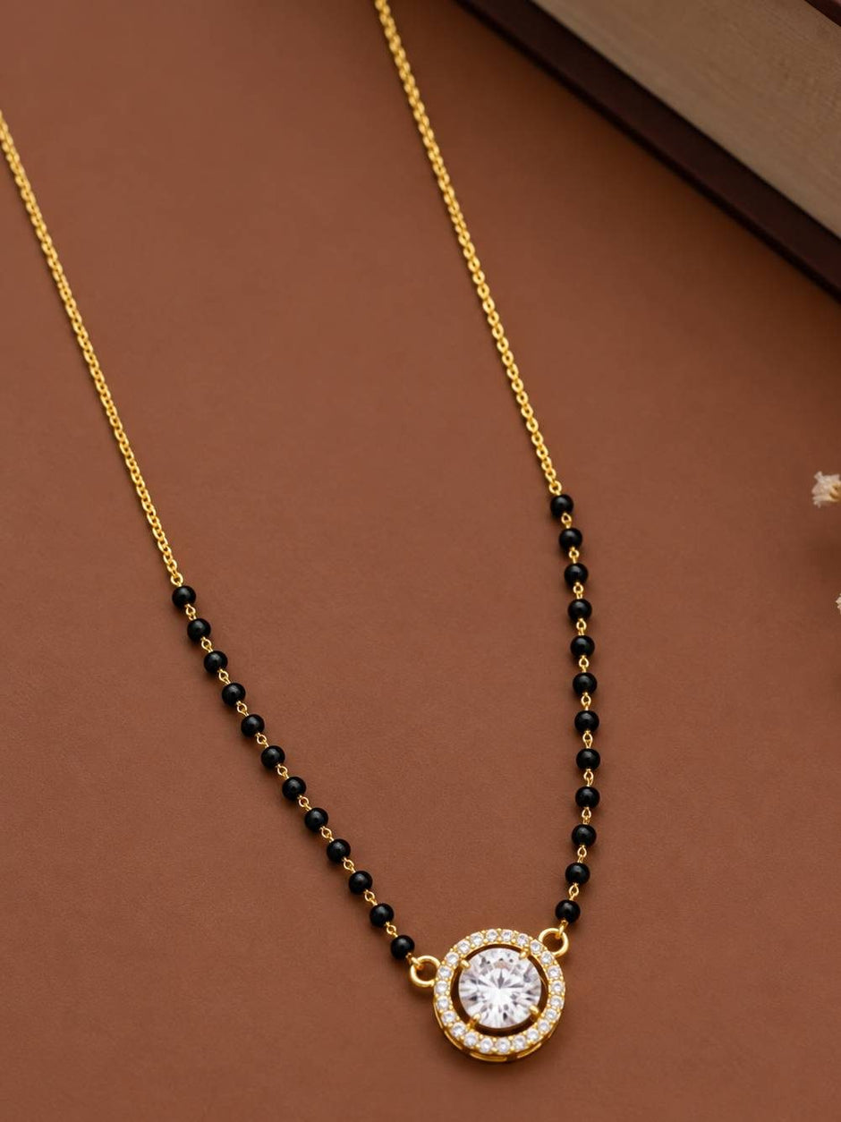 Gold-plated mangalsutra with black beads and circular CZ pendant laid flat on a brown surface