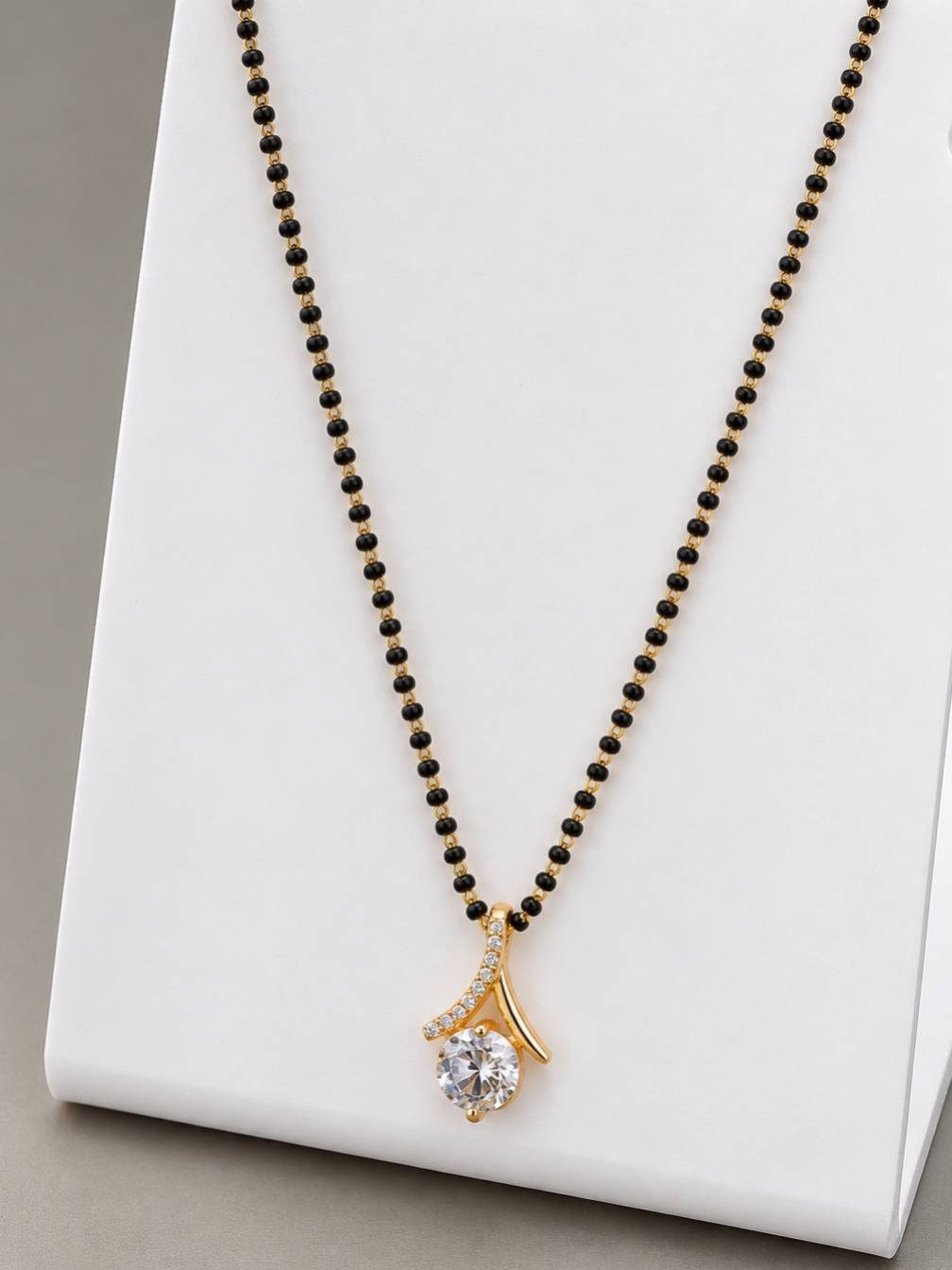 Gold-plated black bead mangalsutra with CZ solitaire pendant displayed on a white jewellery stand