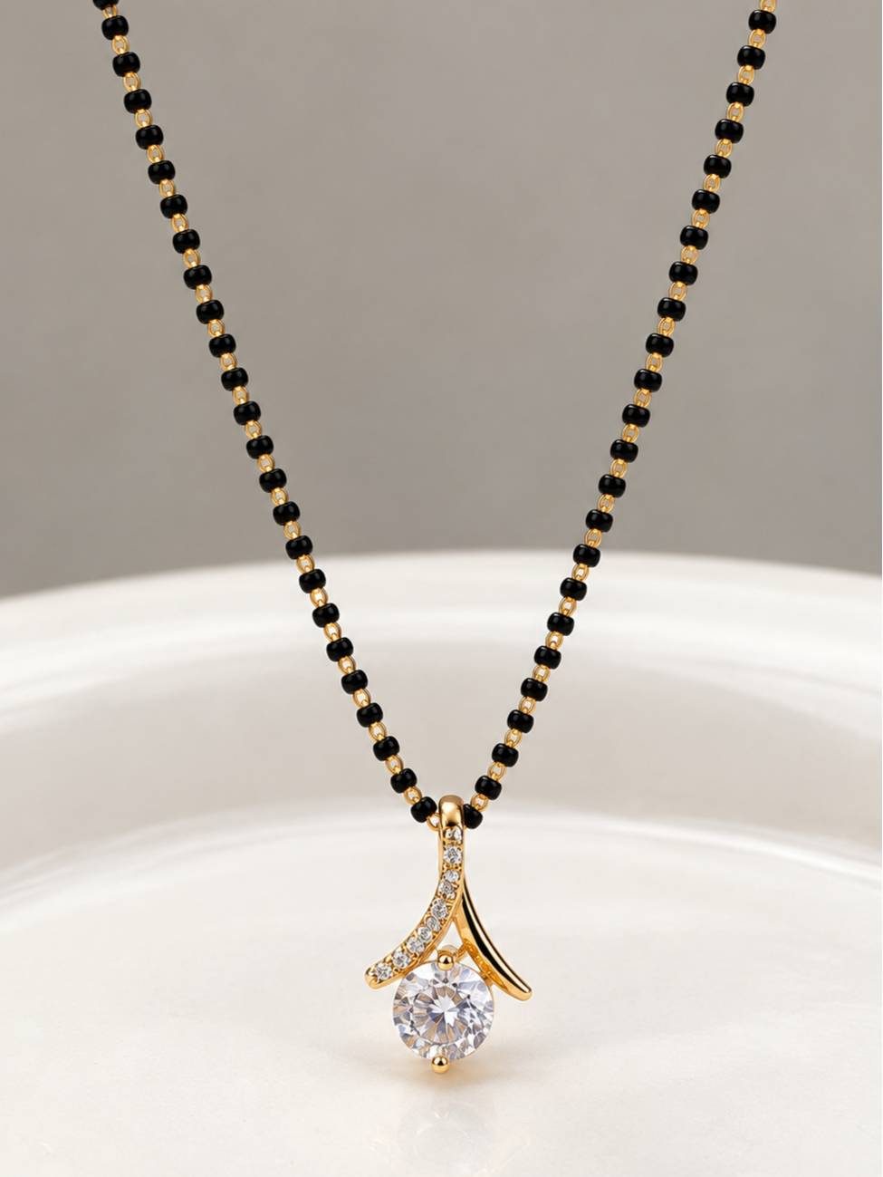 Black bead mangalsutra with gold-plated CZ solitaire pendant laid flat on a white curved surface