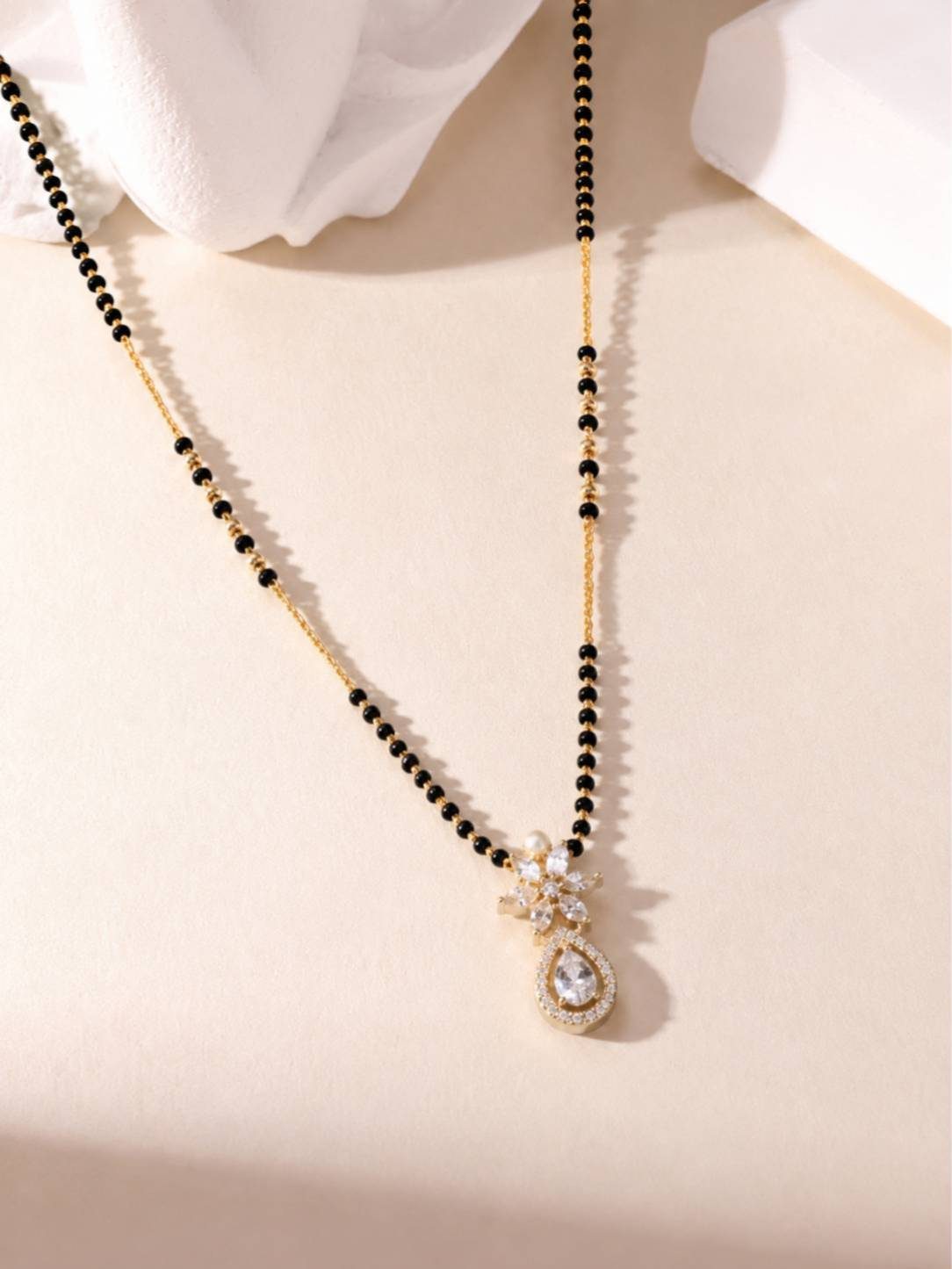 Flat lay of gold-plated mangalsutra with marquise CZ flower and teardrop halo pendant on cream surface