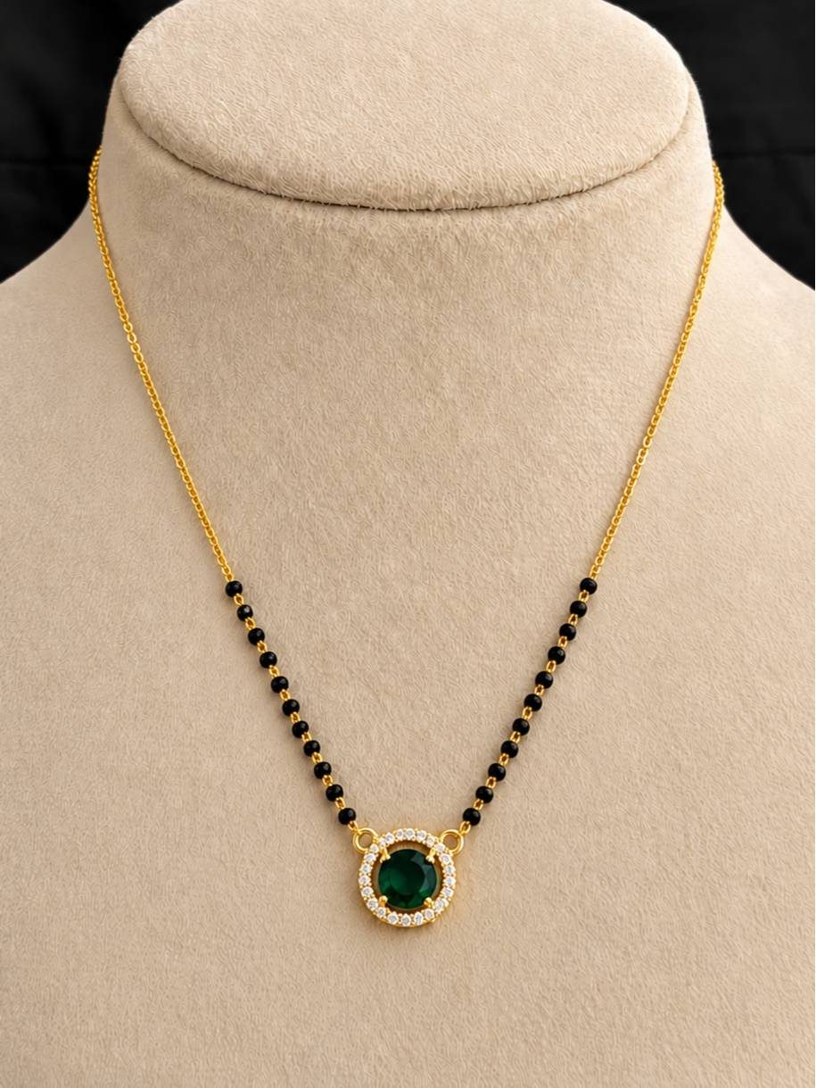 Gold-plated mangalsutra with emerald green CZ halo pendant displayed on a cream jewellery bust
