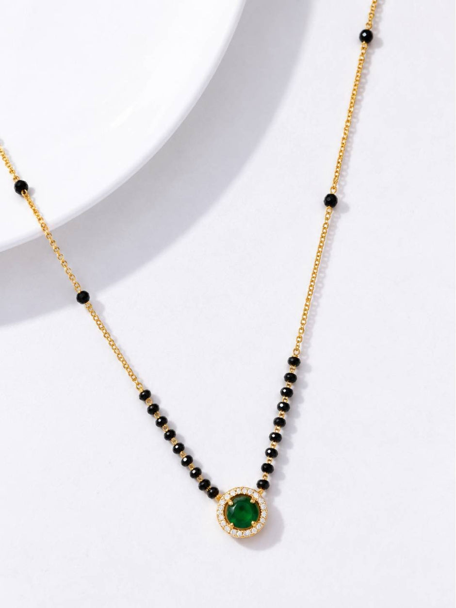 Close-up of emerald CZ halo pendant on gold-toned black bead mangalsutra chain on white surface
