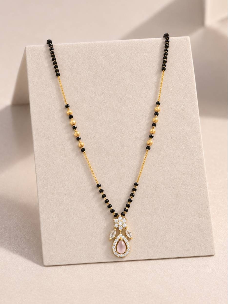 Gold-plated mangalsutra with black beads and rose pink CZ pendant displayed on a cream jewellery stand