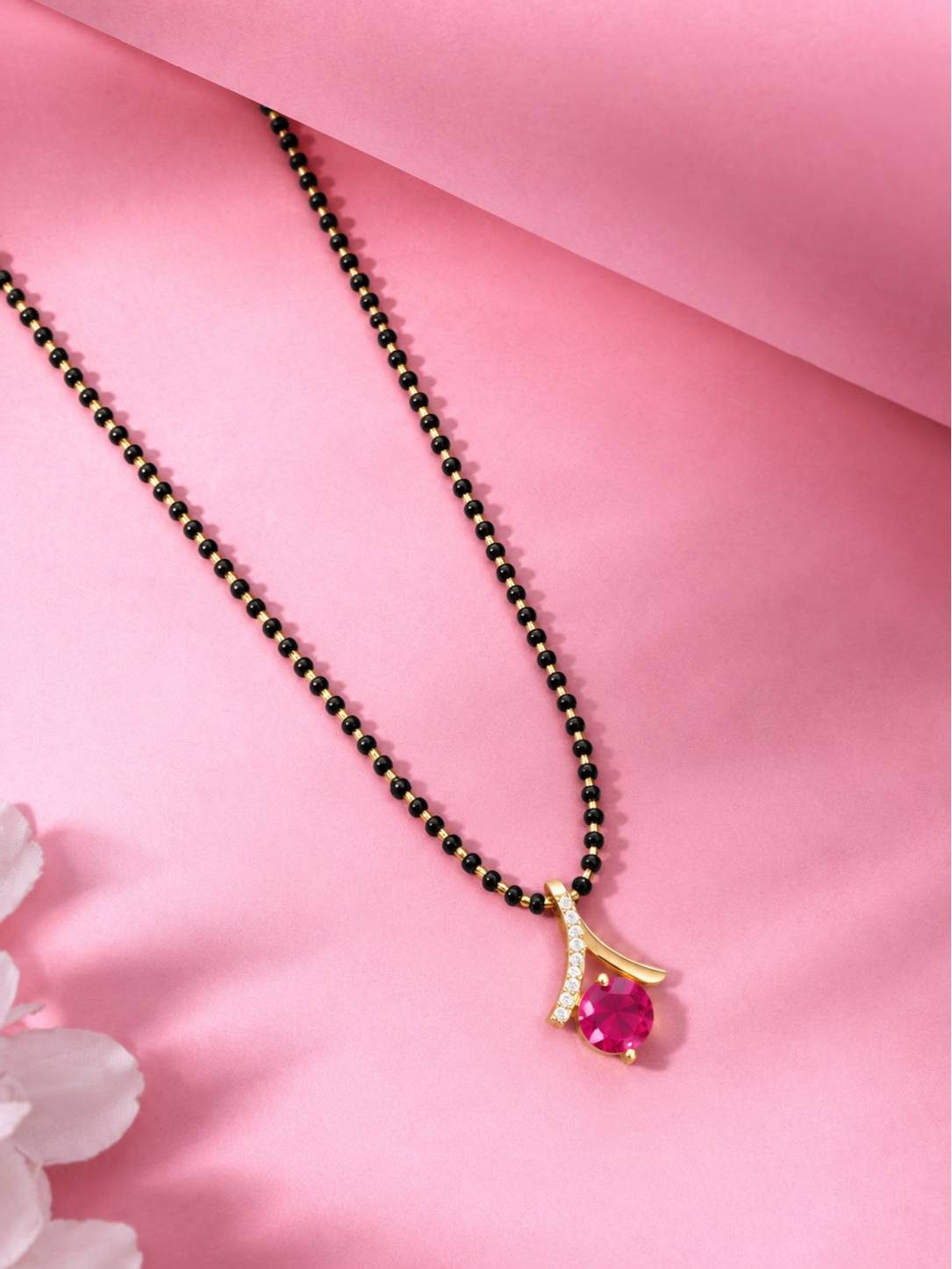 Gold-plated black bead mangalsutra with curved pendant resting on a pink fabric surface beside white flowers