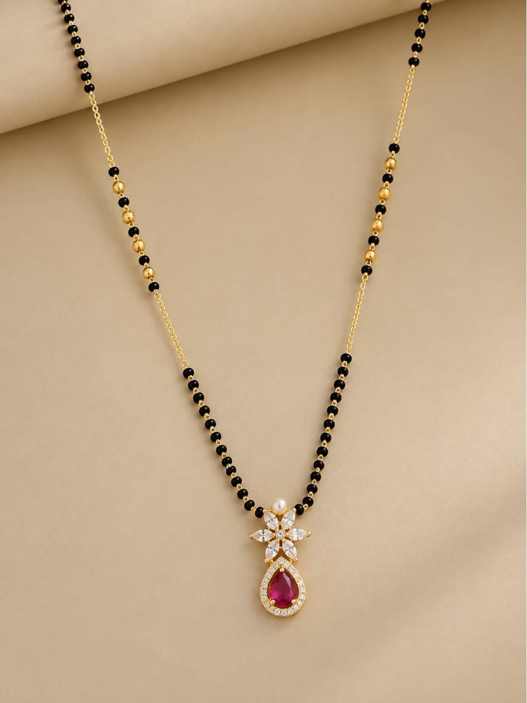 Full-length view of black bead gold mangalsutra with fuchsia CZ drop pendant on beige fabric