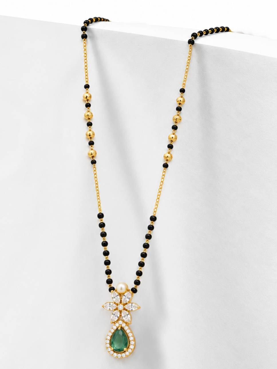 Side-angle view of gold-plated mangalsutra with black beads and emerald CZ pendant against white background