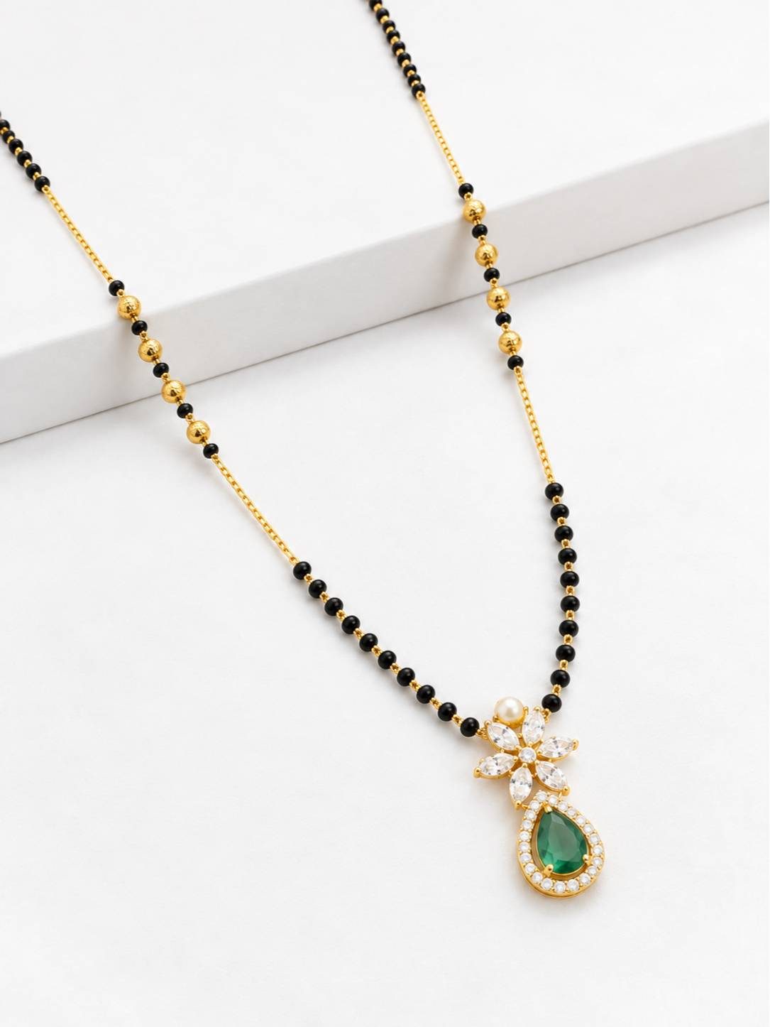 Flat lay of gold-plated black bead mangalsutra with green stone and white CZ pendant on white surface