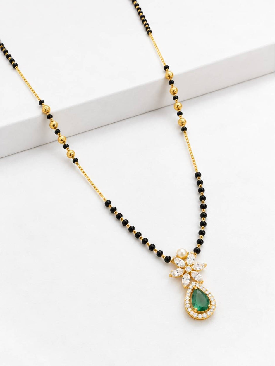 Flat lay of gold-plated black bead mangalsutra with green stone and white CZ pendant on white surface