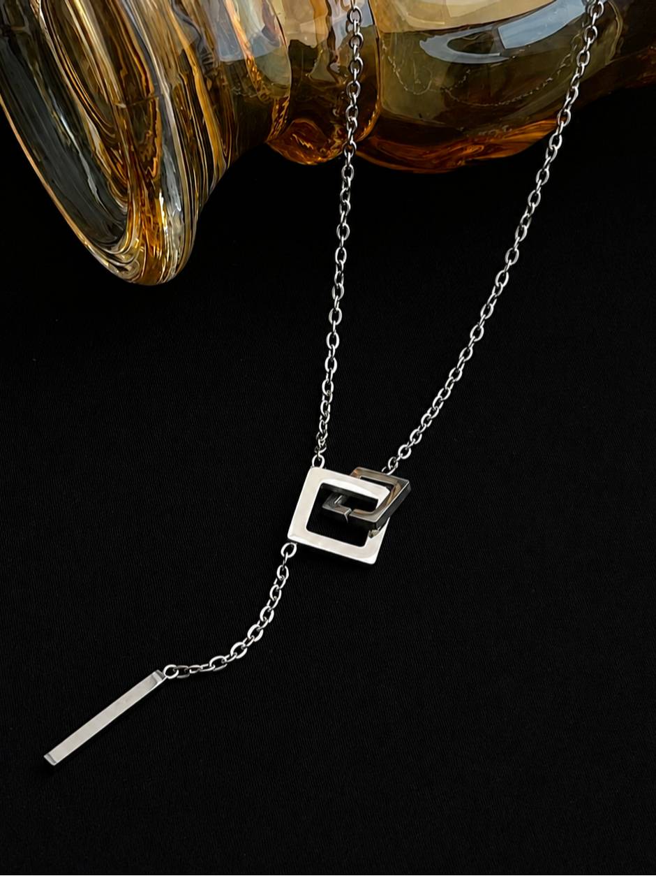 Close-up of interlocked square pendant and bar drop on silver lariat necklace, laid flat beside an amber glass object