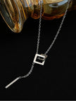Close-up of interlocked square pendant and bar drop on silver lariat necklace, laid flat beside an amber glass object