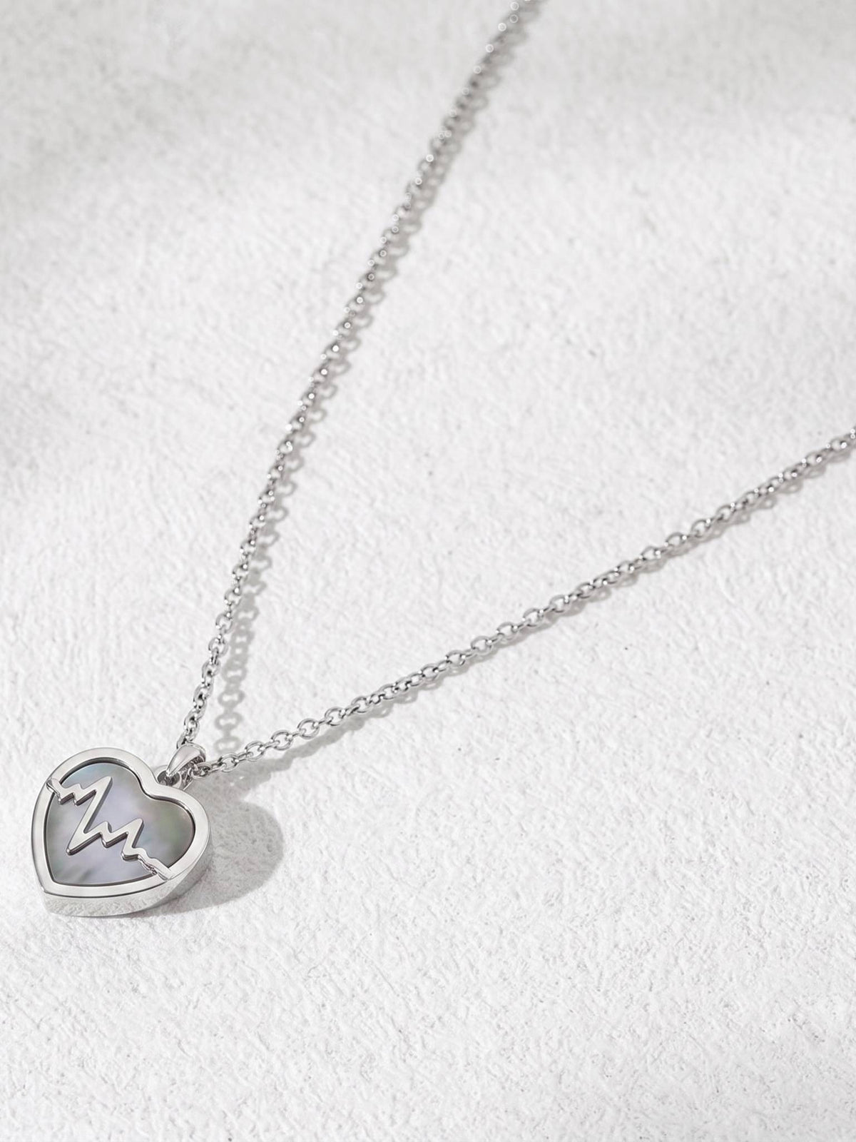 Angled flat-lay of silver heartbeat heart pendant necklace on textured white surface