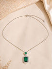 Silver-finish emerald pendant necklace laid flat on a beige surface with satin fabric in the background