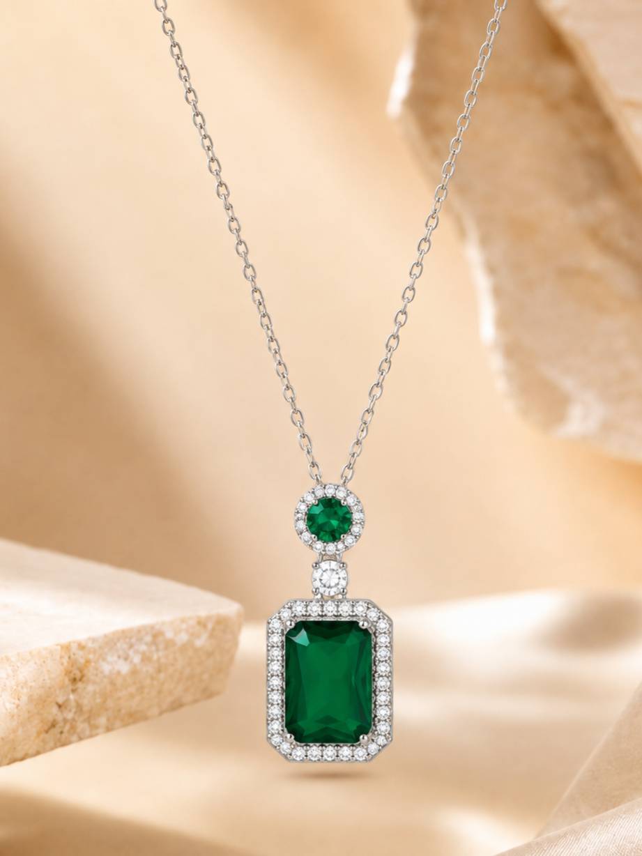 Emerald green rectangular pendant necklace with round CZ halo shown against a soft beige background with stone props