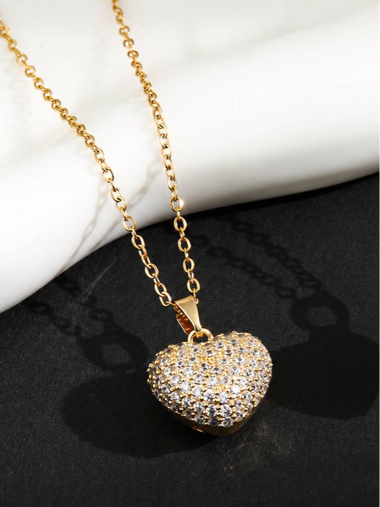 Puffy gold heart pendant with cubic zirconia stones on a cable chain against marble and black surface
