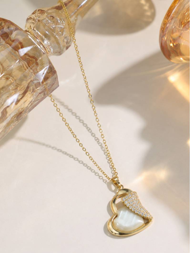 Heart pendant necklace with mother-of-pearl and CZ lying flat on a light surface with warm-toned glass props in background