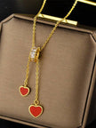 Gold-plated lariat necklace with two red enamel heart charms displayed on a brown jewellery box