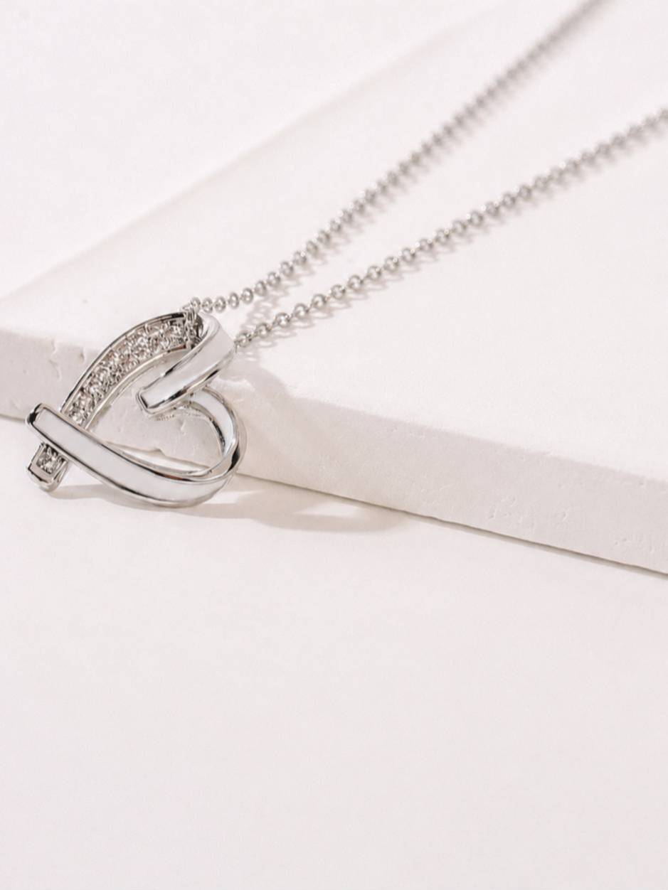 Close-up of rhodium-plated heart pendant with pavé CZ arch resting on a white surface, showing twisted ribbon detail