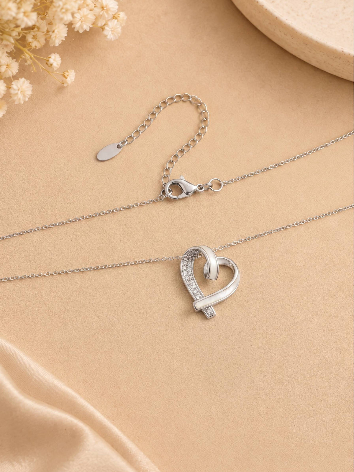Full silver heart pendant necklace laid flat on a beige fabric surface showing lobster clasp, extender chain and oval tag