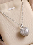 Silver-finish pavé cubic zirconia heart pendant necklace laid on a cream fabric surface showing lobster clasp detail