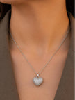 Model wearing silver cubic zirconia heart pendant necklace at collarbone level over a brown blazer