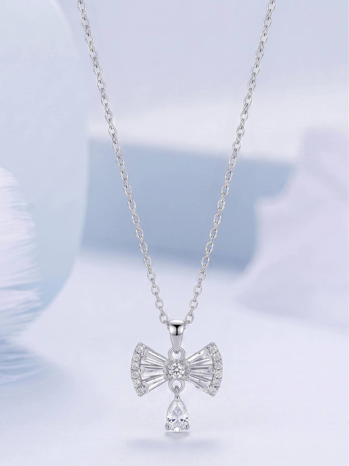 Silver cubic zirconia bow pendant necklace hanging against a soft blue background showing full silhouette