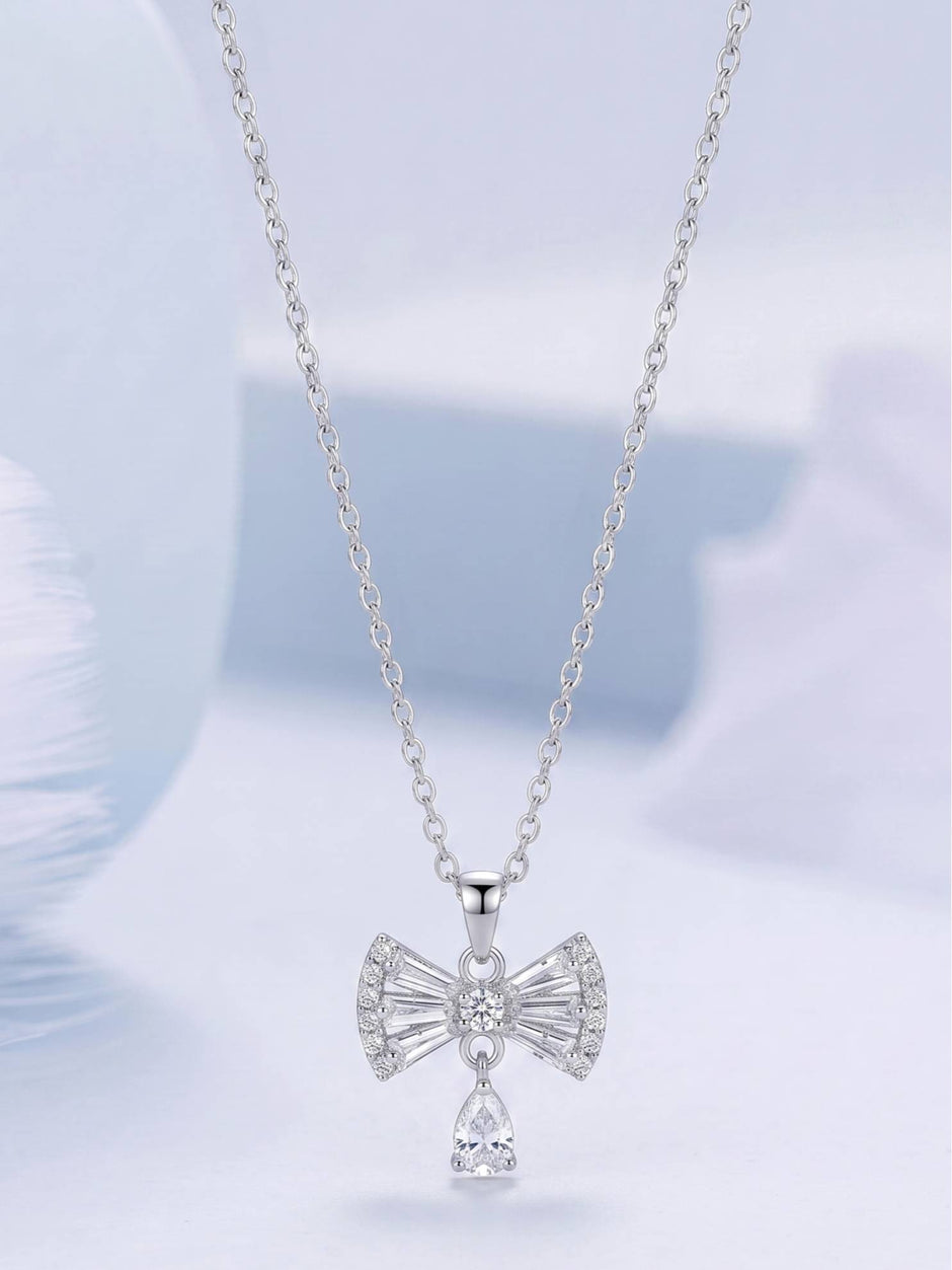 Silver cubic zirconia bow pendant necklace hanging against a soft blue background showing full silhouette