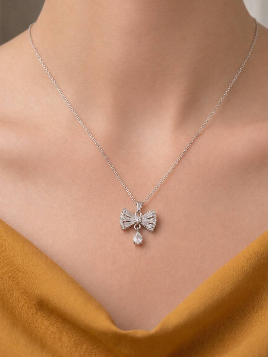 Model wearing silver bow pendant necklace over a mustard yellow satin top at collarbone length