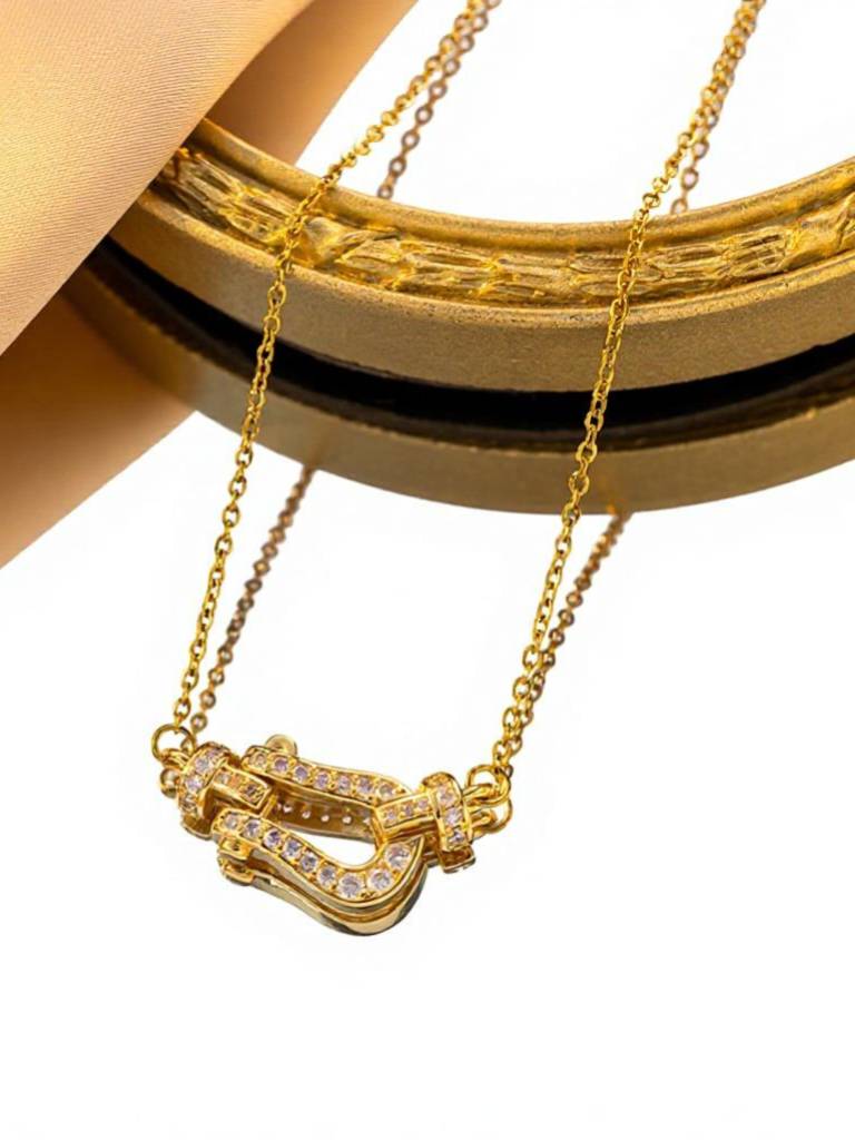 Gold-plated CZ horseshoe clasp necklace hanging against a gold ornate frame on white background