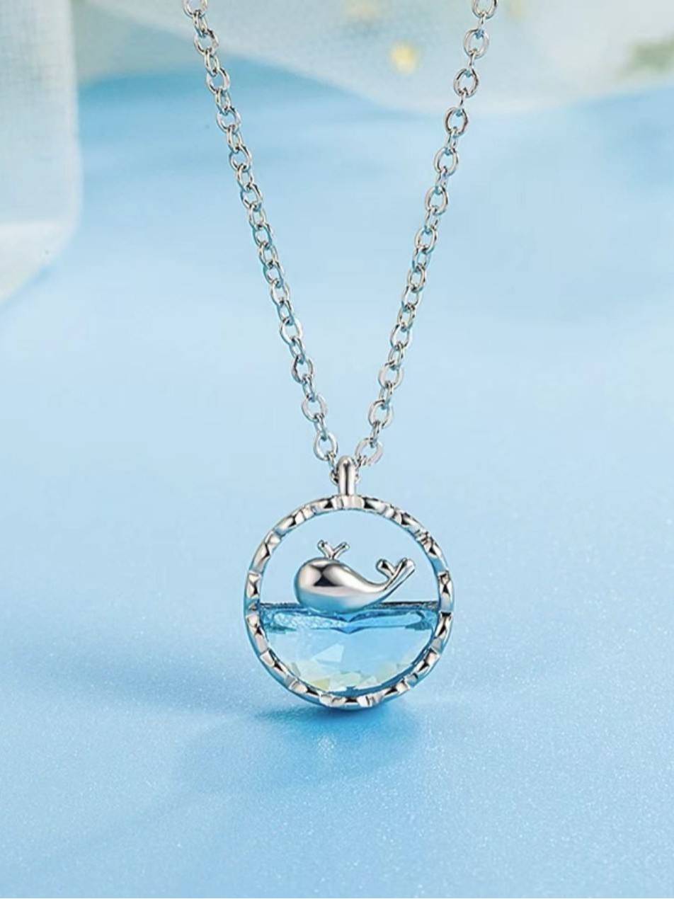 Silver-tone circular pendant necklace with aqua crystal and whale charm on blue background