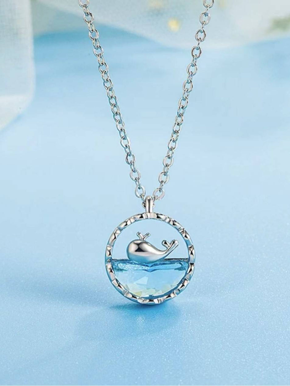 Silver-tone circular pendant necklace with aqua crystal and whale charm on blue background