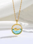 Gold-plated circular pendant with aqua crystal and dolphin detail hanging on cable chain against white fabric