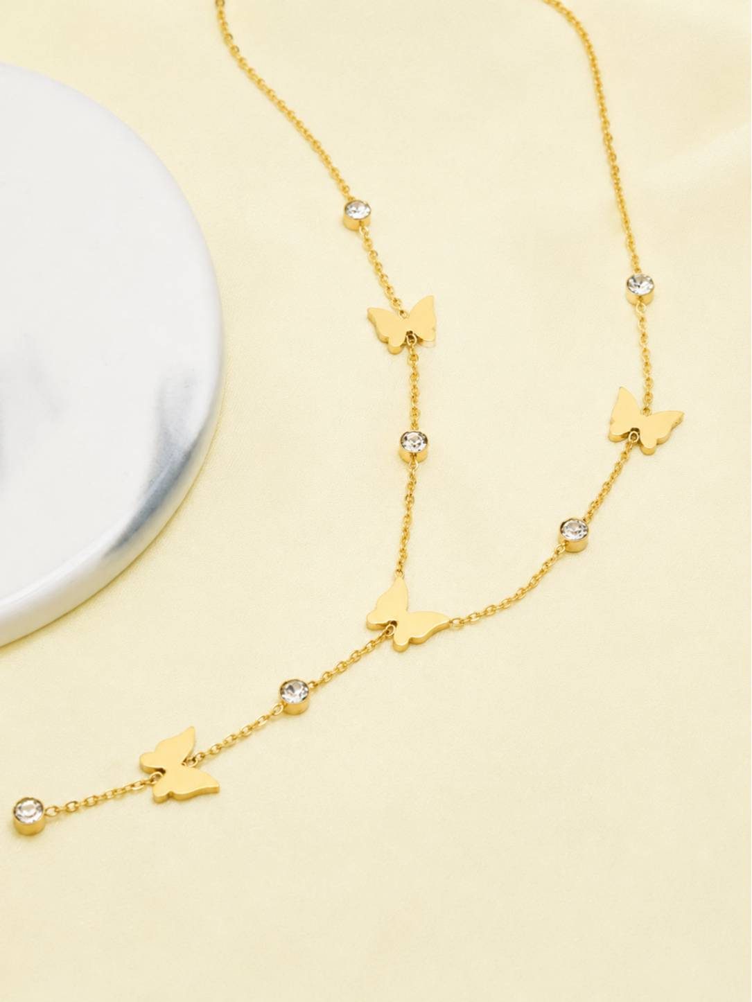 Gold butterfly lariat necklace laid on a cream surface beside a marble disc, highlighting the drop chain with butterfly and crystal detailing