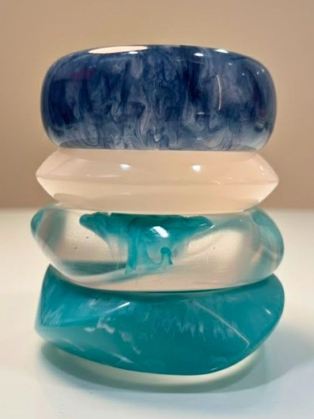 Four stacked marbled resin bangles in navy, milky white, clear teal, and solid turquoise on a white surface
