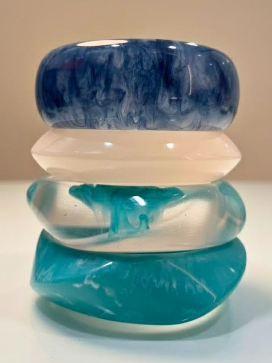 Four stacked marbled resin bangles in navy, milky white, clear teal, and solid turquoise on a white surface