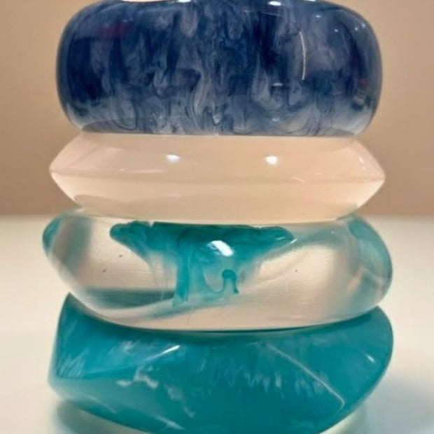 Four stacked marbled resin bangles in navy, milky white, clear teal, and solid turquoise on a white surface