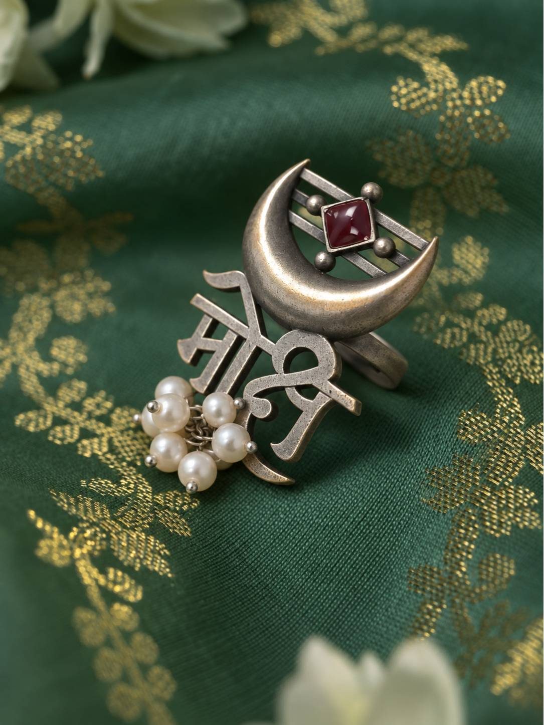 Oxidised silver Moksh script ring with crescent moon, red stone and pearl drops laid flat on green brocade fabric