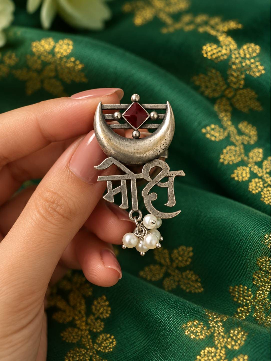 Close-up of Moksh script ring held in hand showing red stone, crescent moon motif and clustered pearl drops against green brocade