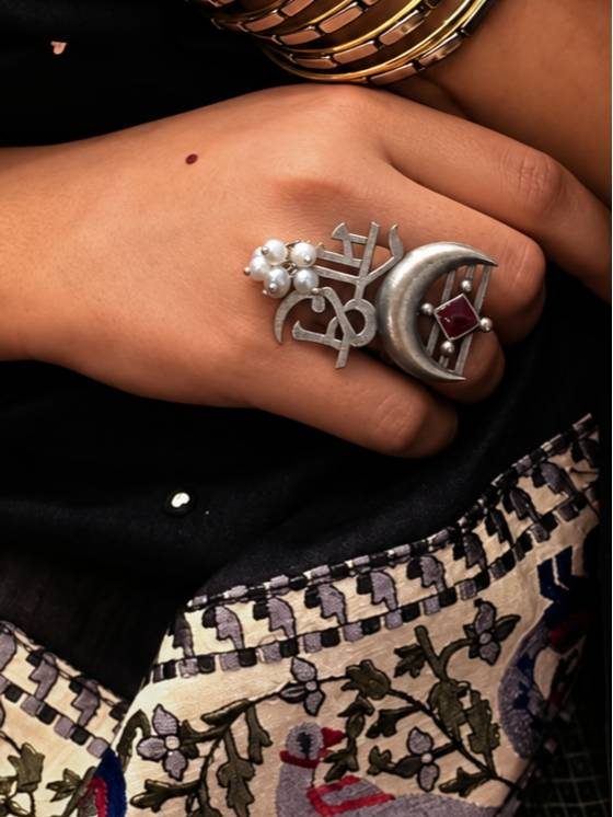 Moksh script ring worn on hand against black embroidered fabric showing pearl drops and crescent moon from front angle