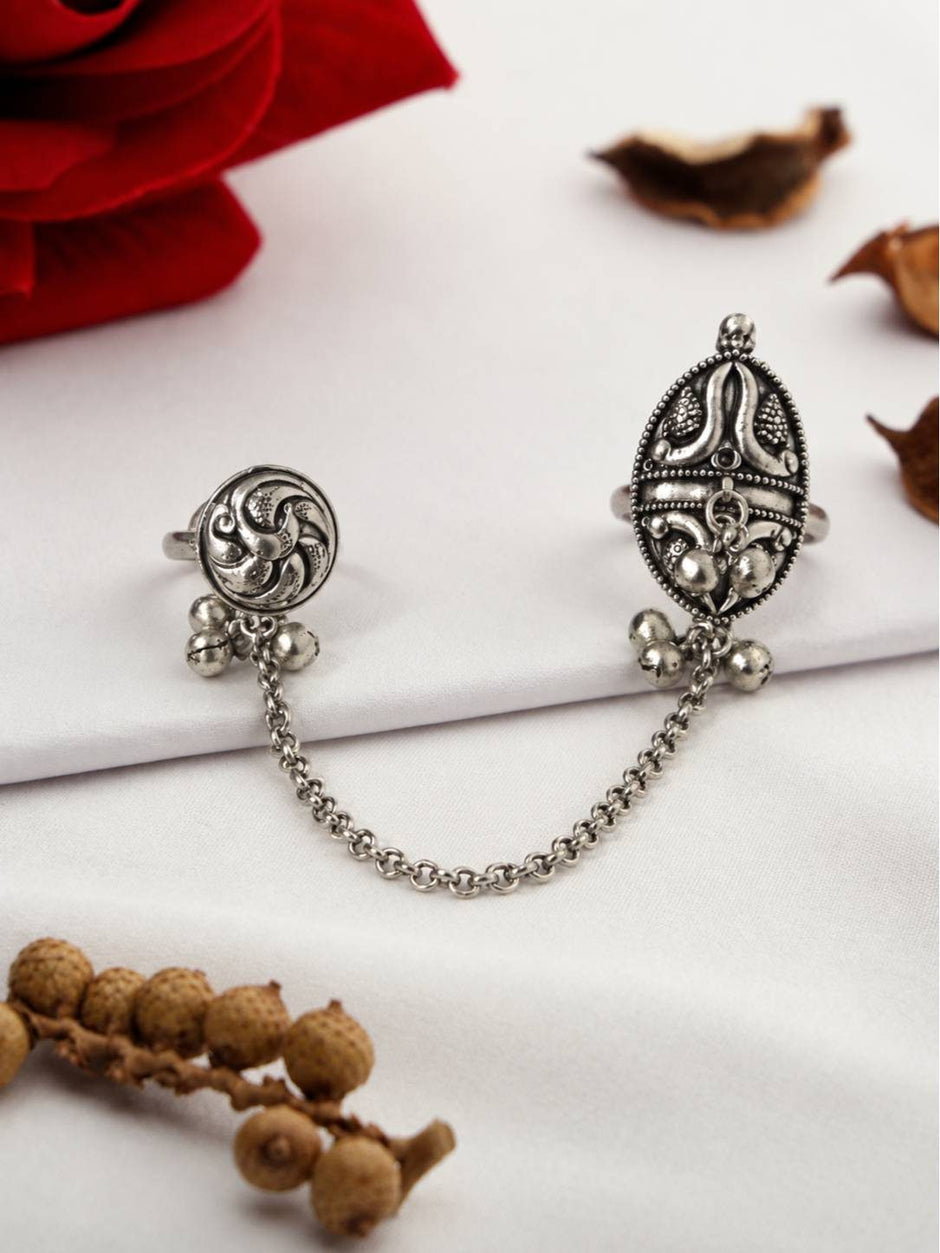 Oxidised silver dual finger ring with oval and round tops, ghungroo bells, and connecting rolo chain laid flat on white fabric