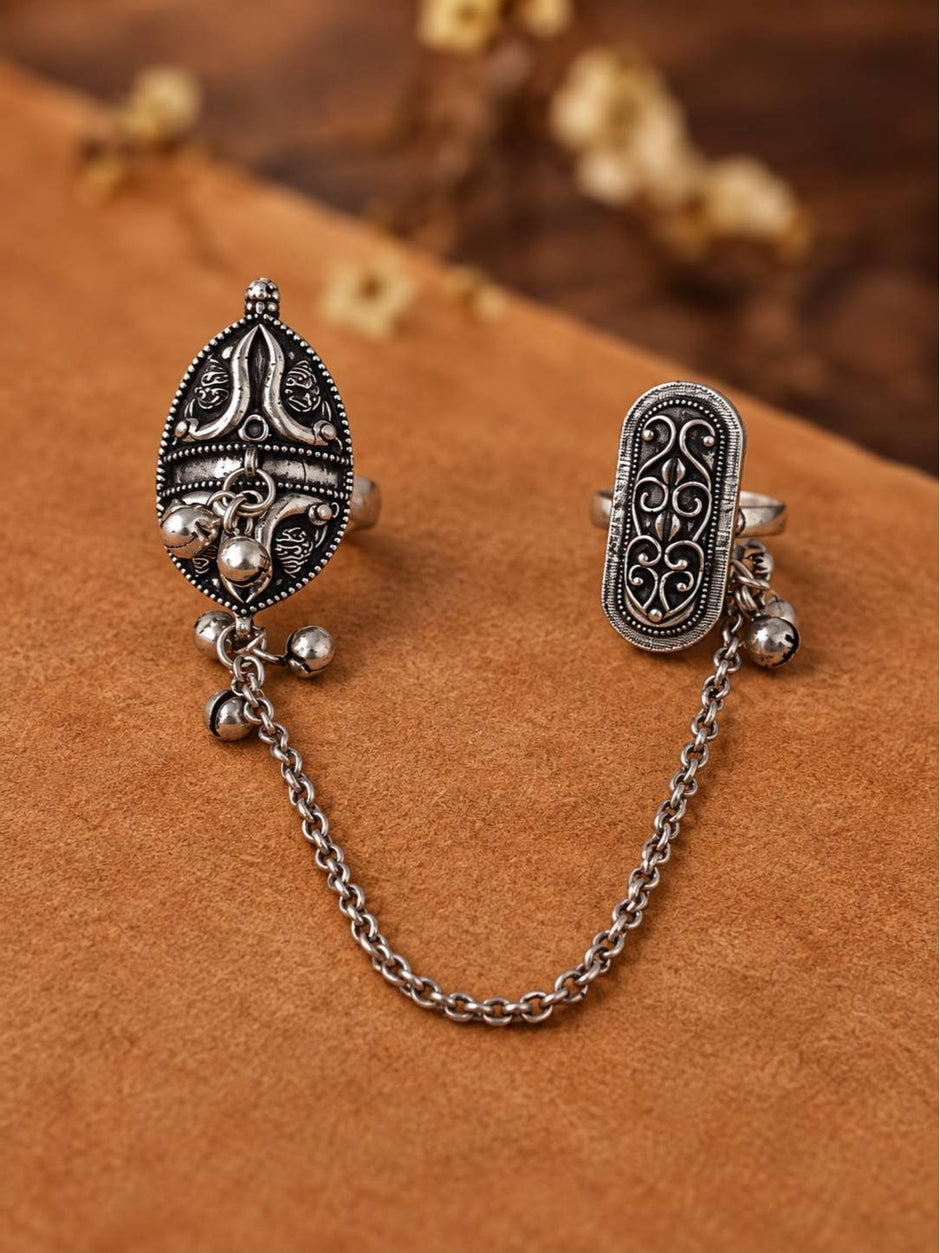 Two oxidised silver engraved rings connected by a rolo chain with ghungroo bells, displayed on a brown suede surface