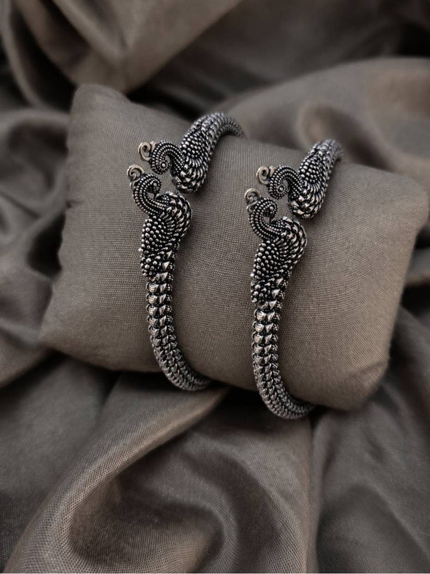 Pair of oxidised silver peacock bangles resting on a grey fabric cushion, showing detailed granule work