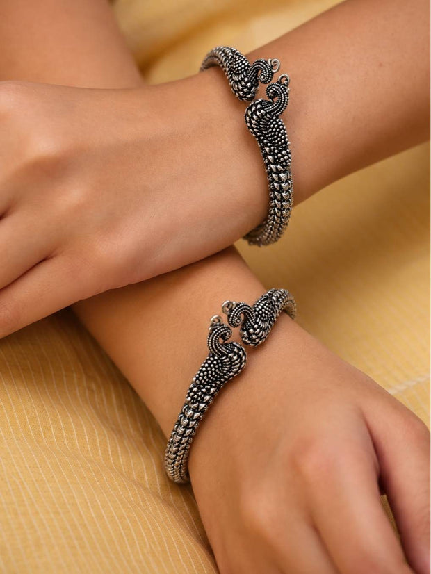 Oxidised silver peacock bangles worn on both wrists of a woman against a yellow fabric background