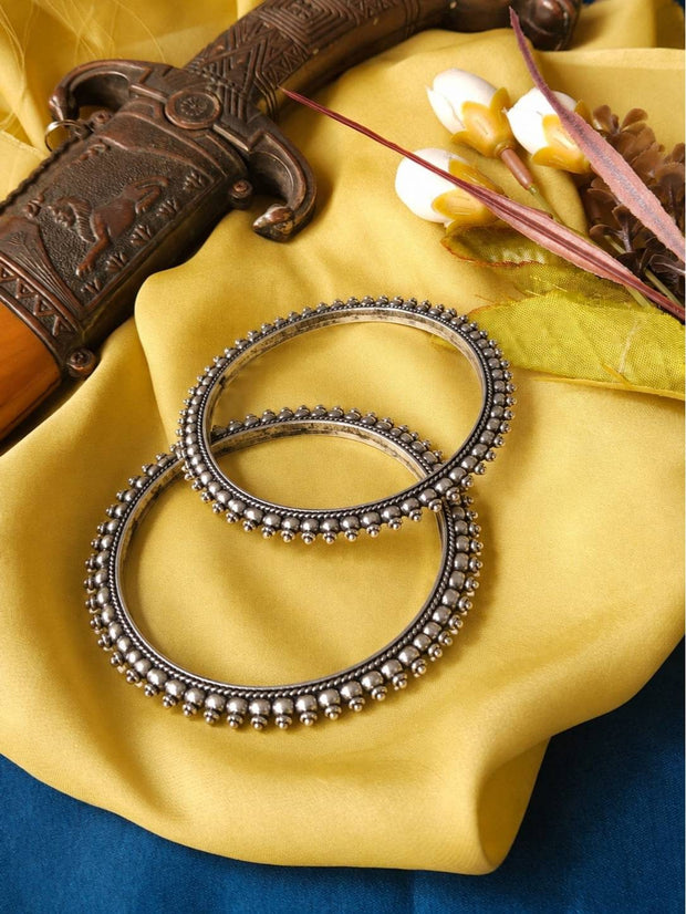 Pair of oxidised silver ghungroo kada bangles with double beaded border resting on yellow fabric beside an antique dagger prop