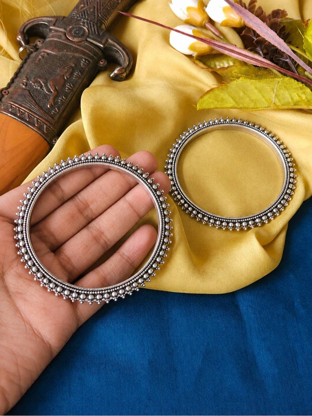 Hand holding one oxidised silver beaded bangle flat against yellow fabric, second bangle resting beside it showing full circular shape