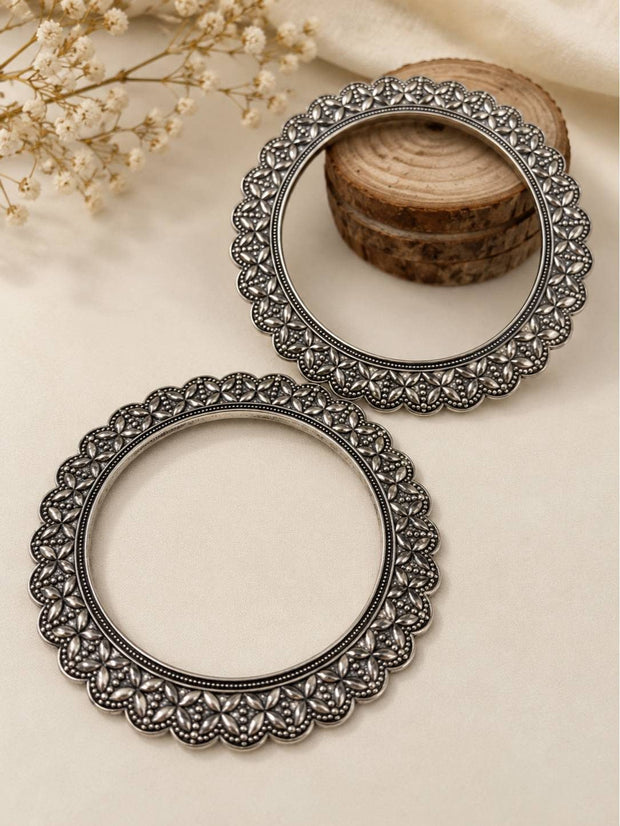 Pair of oxidised silver floral kada bangles laid flat on a cream surface with dried flowers and wooden slices