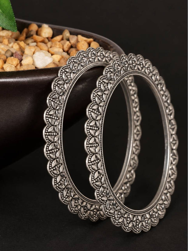 Two oxidised silver floral bangles leaning upright against a dark bowl with pebbles in the background
