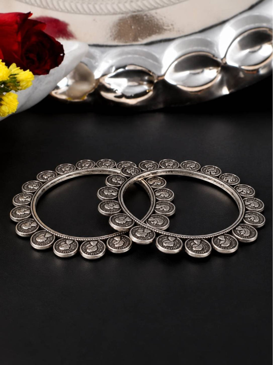 Pair of oxidised silver coin bangles lying flat on a dark surface with flowers and a silver tray in the background